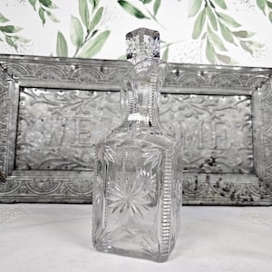 May include: A clear glass decanter featuring a square base and a matching square stopper. The body of the decanter is decorated with embossed starburst patterns on the sides and vertical ribbed detailing along the edges and the neck.