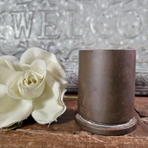 May include: A cylindrical brown metal object with a flat base and a smooth surface, positioned next to a single white artificial rose with layered petals. The metal item has a weathered appearance with subtle variations in its dark brown tone.