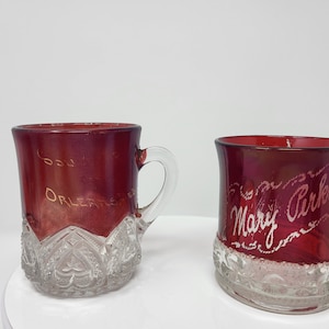 May include: Two vintage ruby red glass cups with clear glass bases. One cup has the words "New Orleans" in gold lettering. The other cup has the words "Mary Perkins" in white lettering.