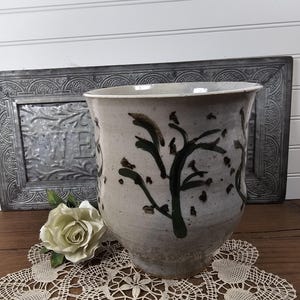 May include: A ceramic planter with a wide, flared rim and a tapered base. The exterior features a light gray, speckled glaze decorated with dark brown, hand-painted abstract tree or branch motifs encircling the body of the vessel.