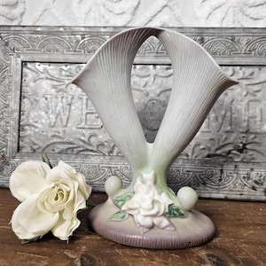 May include: A vintage, off-white ceramic vase with a unique, open design. The vase features two upward-curving, petal-like forms, a floral base, and two spherical accents. The color palette includes soft shades of white, lavender, and green.