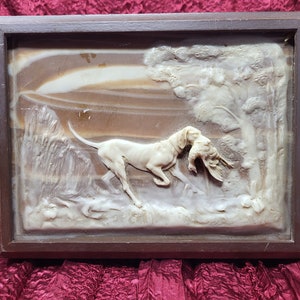 May include: A framed relief sculpture depicting a hunting scene. A cream-colored dog carries a bird in its mouth, set against a landscape with trees and a mountain. The artwork is encased in a dark brown wooden frame.