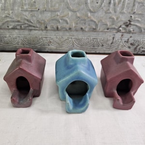 May include: Three ceramic birdhouse-shaped incense holders in shades of dusty rose and blue. Each has a chimney, a round opening, and a small ramp. The birdhouses are arranged on a white surface.