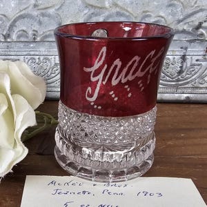 May include: A vintage red glass tumbler with the name "Grace" etched in white script. The top portion is red, while the base features a diamond-cut pattern. The glass is approximately 5 oz.