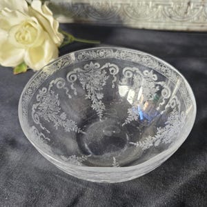 May include: A clear glass bowl with etched floral and scroll designs. The bowl has a rounded shape and a decorative border. The intricate patterns are visible against the clear glass, creating a delicate and elegant appearance. A white flower is in the background.
