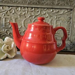 May include: A vibrant red ceramic teapot with a matching lid and a curved handle. The teapot has a glossy finish and a classic design, perfect for serving tea. The teapot is approximately 8 inches tall.