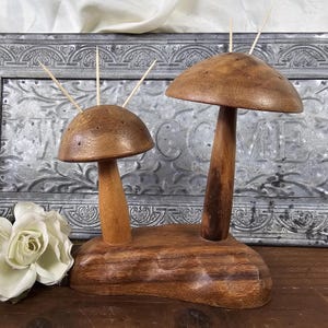 May include: Wooden mushroom toothpick holders. The holders are made of wood and feature a large and small mushroom design. The mushrooms have a brown finish and are attached to a wooden base. Toothpicks are inserted into the top of the mushrooms.