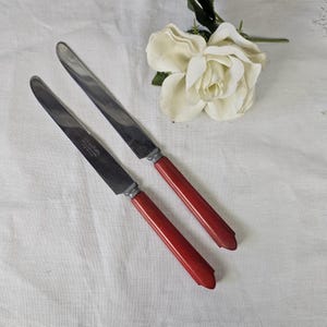 May include: Two vintage stainless steel knives with red handles. The blades are silver and the handles are a deep red color. The knives are placed on a white surface, with a white flower in the background.