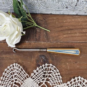 May include: A vintage crochet hook with a silver metal shaft and a decorative handle featuring a cream, blue, and gold design. The hook is positioned on a wooden surface, with a white rose and lace doily in the background.