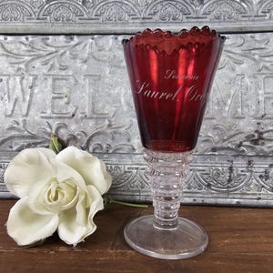 May include: A vintage ruby red glass souvenir goblet with a clear, ribbed stem and base. The goblet has the words "Souvenir Laurel, Ore" etched on the front. A white flower rests near the glass.