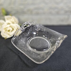 May include: A clear, square-shaped glass dish with an oval center and etched floral designs. The edges are scalloped, and the dish is set against a dark background. The dish is likely vintage or antique.