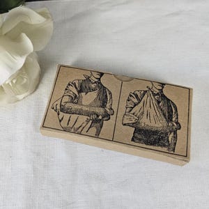 May include: A small, rectangular, vintage-style box with illustrations depicting arm sling techniques. The box is a light brown color with black and white drawings. The illustrations show how to use a sling for an injured arm.