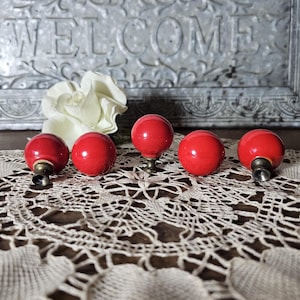 May include: Five red, round cabinet knobs with brass hardware are arranged on a cream-colored lace doily. A white flower and a metal sign that says "WELCOME" are in the background. The knobs are a vibrant, glossy red.