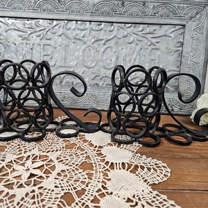 May include: A black metal welcome sign with a decorative design of interlocking circles. The sign is sitting on a white lace doily on a wooden surface.