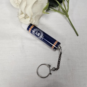 May include: A vintage blue and orange New York World's Fair 1939 souvenir keychain. The cylindrical keychain has a silver chain and key ring. A white rose and green stem are in the background.