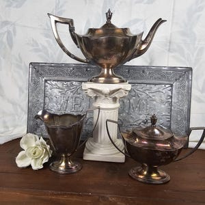 May include: A set of three silver-plated serving pieces, including a teapot, creamer, and sugar bowl. The pieces are arranged on a white pedestal with a gray metal background.
