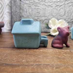 May include: A light blue ceramic house-shaped planter with a small handle. A small, purple-to-red gradient pig figurine sits nearby. A light blue house-shaped object with a circular opening is also visible. The items are on a wooden surface.