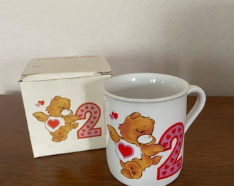 May include: White ceramic mug with a brown bear graphic, a pink number 2, and red hearts. The mug has a handle and is next to a matching box with the same design. The mug is likely for a child's second birthday.