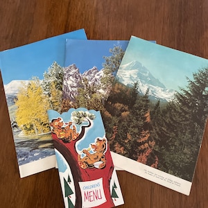 May include: A vintage children's menu with a red background and the words "Children's Menu" in white. The menu features a cartoon illustration of two bears sitting in a tree, eating from bowls. The menu is surrounded by three vintage postcards with images of mountains and forests.
