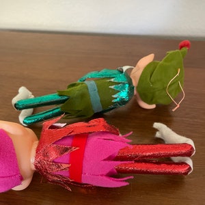 May include: Two felt Christmas elves, one in green with a red hat and one in pink with a red and gold hat.