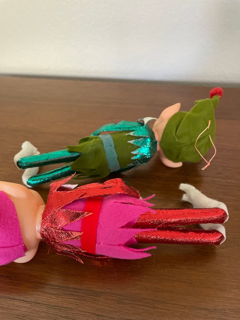 May include: Two felt Christmas elves, one in green with a red hat and one in pink with a red and gold hat.