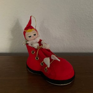 May include: A red felt Christmas ornament shaped like a boot with a small elf doll sitting inside. The elf has blonde hair, a red hat, and a white fur collar.