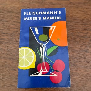May include: A vintage cocktail mixer's manual titled "FLEISCHMANN'S MIXER'S MANUAL" on a blue background. The cover features a martini glass with an olive, an orange slice, a lemon slice, and cherries.
