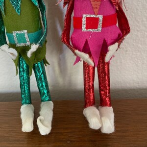 May include: Two Christmas elf ornaments, one with a green and teal outfit and the other with a pink and red outfit. Both have white felt boots.