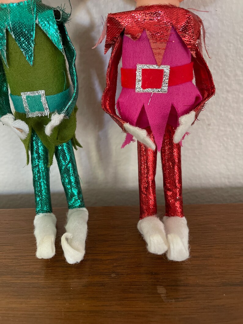 May include: Two Christmas elf ornaments, one with a green and teal outfit and the other with a pink and red outfit. Both have white felt boots.