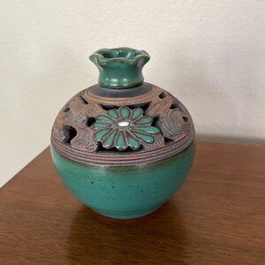 May include: A round ceramic incense burner featuring a deep teal glazed base and a dark brown upper section with intricate cut-out patterns. The top is adorned with a sculpted teal flower motif and a small, flared opening at the center.