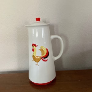 May include: White ceramic pitcher with a red base and lid, featuring a colorful rooster illustration. The pitcher has a handle and a slightly tapered shape, perfect for serving beverages.