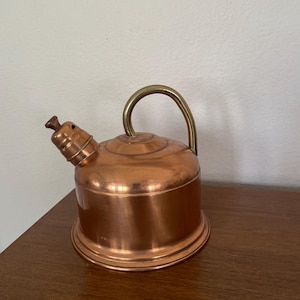 May include: A copper kettle with a cylindrical body, a wide base, and a curved brass handle. The kettle features a short, angled spout topped with a small, dark brown wooden knob. The metallic surface shows a polished finish with subtle reflections.