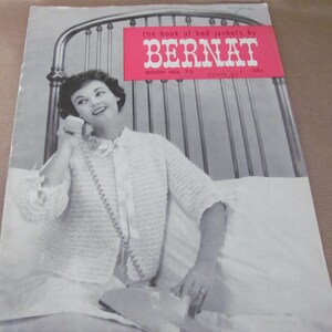 May include: A vintage Bernat knitting pattern book featuring a woman wearing a white crocheted bed jacket. The book cover has a pink title that reads "the book of bed jackets by BERNAT" and "BOOK NO. 72".