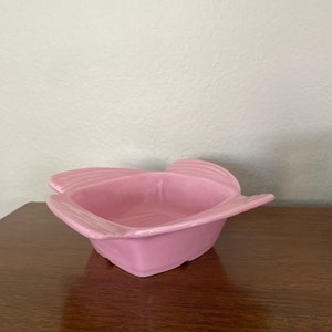 May include: A small, pink ceramic dish with a square shape and rounded edges. The dish has a slightly textured surface and is perfect for serving small snacks or condiments.