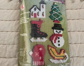 May include: A set of six plastic canvas ornaments for Christmas. The ornaments include a church, a Christmas tree, a boot, a snowman, a mitten, and a sleigh. The package says "Set of 6 Festive Plastic Canvas Ornaments" and "Easy! Fun to Do! A Family Project".