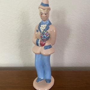 May include: A ceramic figurine of a person wearing a pink hat, blue jacket, and blue pants. The person is holding a bouquet of flowers.