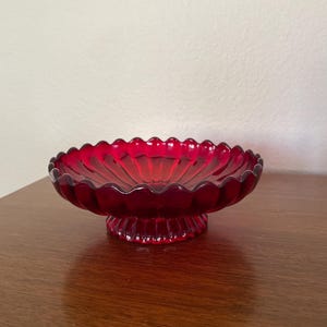 May include: A ruby red glass pedestal bowl with a scalloped rim. The bowl sits on a short, fluted base. The glass is transparent, allowing light to pass through. The bowl is likely vintage or antique.