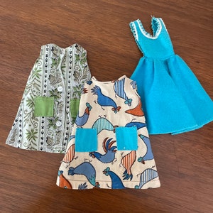 May include: Three doll dresses. One is green and white with a floral pattern, one is cream with a blue and orange rooster pattern, and one is solid blue.