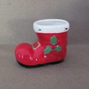 May include: A ceramic Christmas boot-shaped container. The boot is primarily red with a white cuff and black sole. Green holly leaves and berries decorate the side. A gold buckle detail is present. The container is designed to hold small items.