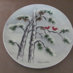 May include: A white ceramic plate with a hand-painted design of two red birds perched on a snow-covered pine tree branch. The plate is signed "Hashimura" in the lower right corner.