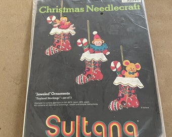 May include: A set of three Christmas stocking ornaments with a red, white, and green color scheme. The ornaments are decorated with beads, sequins, and other embellishments. The ornaments are packaged in a clear plastic bag with a red and white label that reads "Sultana The Quality Line".