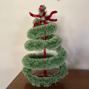 May include: A small, green spiral Christmas tree with a red base and stem. The tree is decorated with a red bow and a silver bell at the top. The tree is sitting on a brown surface.