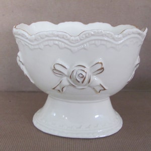 Puede incluir: Taz&oacute;n de cer&aacute;mica blanco con base de pedestal y detalles dorados. El taz&oacute;n presenta un dise&ntilde;o en relieve de una rosa con un lazo.