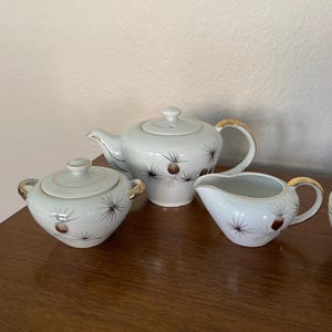May include: A white ceramic tea set with a teapot, sugar bowl, and creamer. The set features a brown and black starburst design and gold trim on the handles and rims. The teapot has a lid, and the sugar bowl has a matching lid.