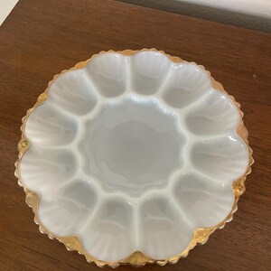May include: A vintage white and gold egg serving platter. The platter has a scalloped edge with gold trim and twelve egg-shaped compartments. The center is a smooth, round surface. The platter is made of glass.