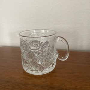 May include: A clear glass mug with a raised design of Batman. The mug has a handle on the right side.