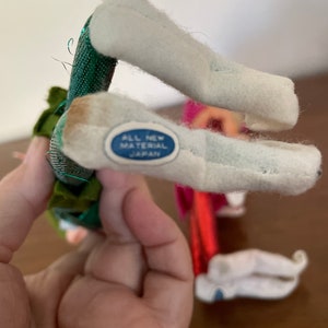 May include: Two felt Christmas elf figures with green and red outfits. The elf on the left has a green outfit with gold glitter and a white hat. The elf on the right has a red outfit with white trim and a white hat. The elf on the left has a label on the bottom of its foot that reads "All New Material Japan".