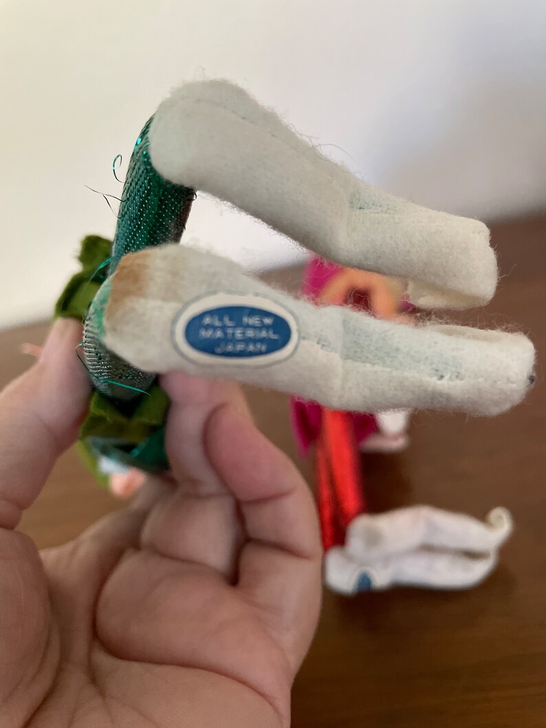 May include: Two felt Christmas elf figures with green and red outfits. The elf on the left has a green outfit with gold glitter and a white hat. The elf on the right has a red outfit with white trim and a white hat. The elf on the left has a label on the bottom of its foot that reads "All New Material Japan".
