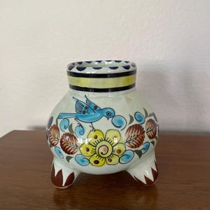 May include: A white ceramic vase with three legs and a blue bird and flower design. The vase has a yellow, black, and blue rim.
