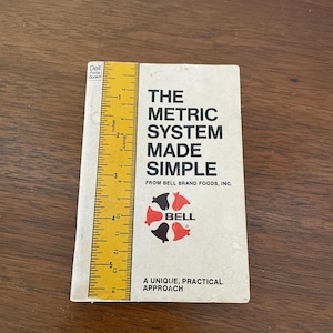 May include: A vintage paperback book titled "The Metric System Made Simple" by Bell Brand Foods, Inc. The cover is off-white with black text and a red and black logo. A yellow ruler is placed next to the book, showing inches.